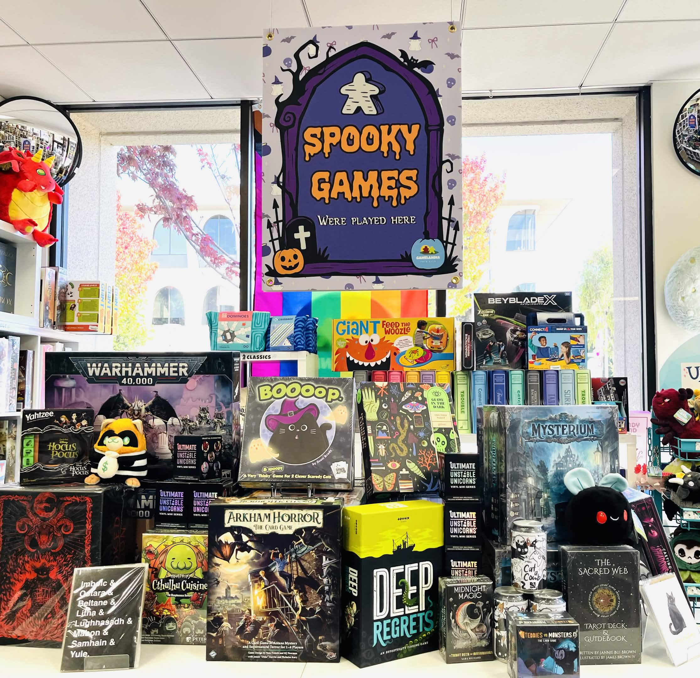A display of spooky-themed board games under a purple Spooky Games sign, with various colorful boxes, plush toys, and decorations on shelves inside a bright, cheerful game store.