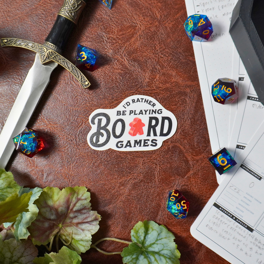 Sword, colorful dice, papers, and an Id Rather Be Playing Board Games sticker on leather surface.