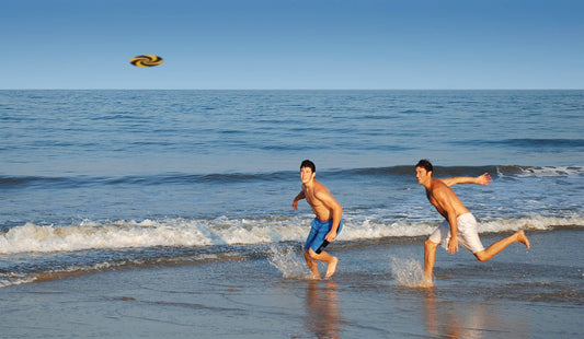 Two men throw an Indoor MayaFlya (Pocket Disc) on a beach.