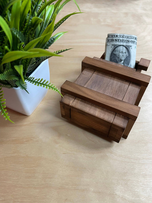 The True Genius Sorcerers Box contains a rolled dollar bill, shown beside a potted plant on a wooden surface.