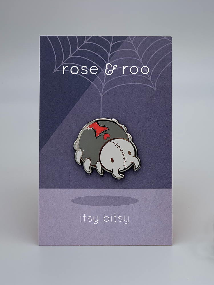 The Itsy Bitsy Enamel Pin shows a stitched zombie black widow spider on a backing card with text and web details.