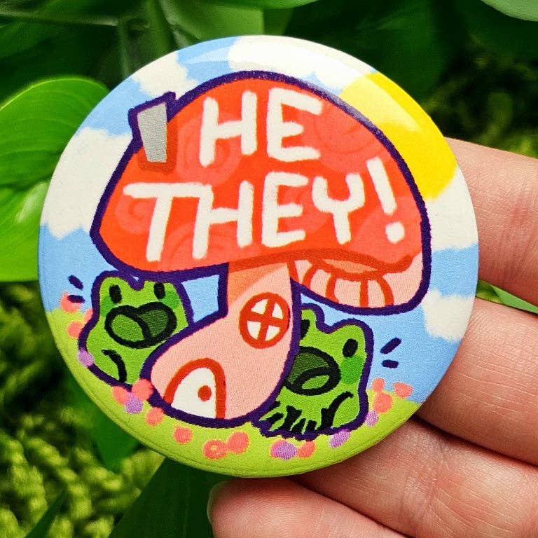 Pronoun Frog Mushroom Large Button with two frogs, a mushroom, and HE THEY in front of green leaves.