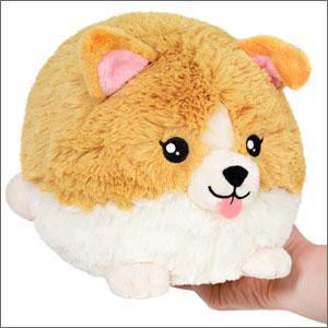 A hand holds a Baby Corgi 10 Mini Squishable plush with big eyes and a pink tongue.