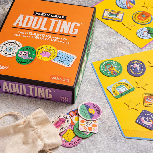 Adulting Party Game box with achievement tokens and scorecards on a table.