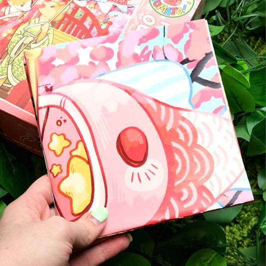 Kimono Kitties 500-piece puzzle box held in hand, featuring koi fish, stars, and green leaves in cartoon style.