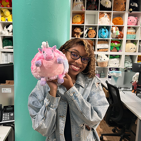Woman in glasses holds a Cotton Candy Baby Unicorn Mini Squishable; plush toys visible on shelves in the background.