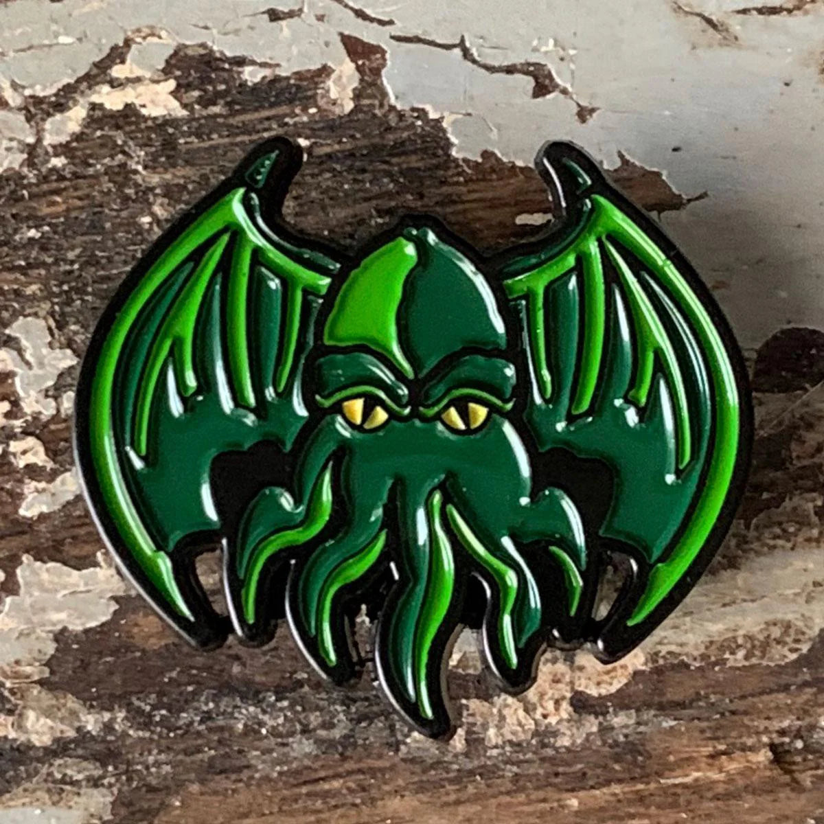 Green Cthulhu enamel pin with wings, yellow eyes, tentacles, and a textured background.