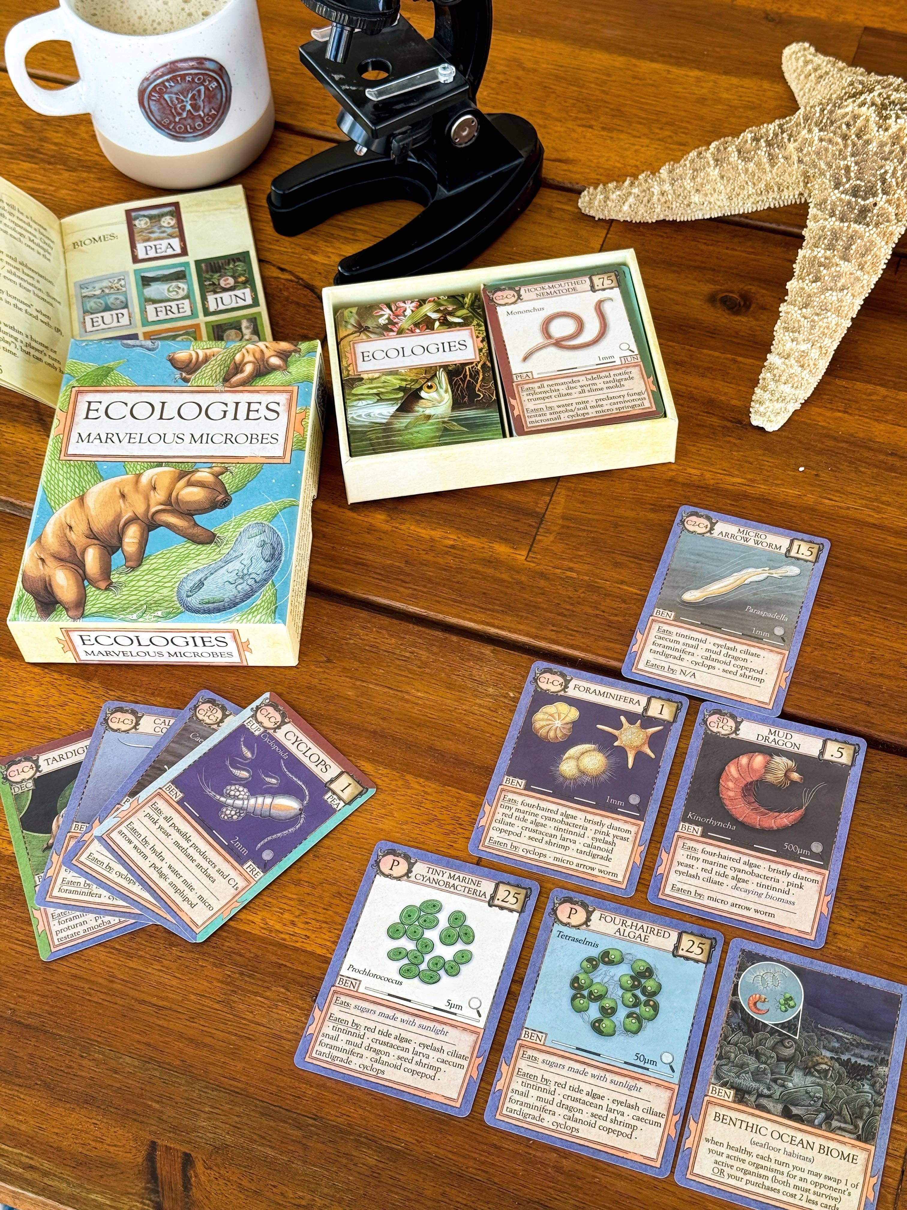Ecologies: Marvelous Microbes card game with cards and rulebook arranged on a wooden table.