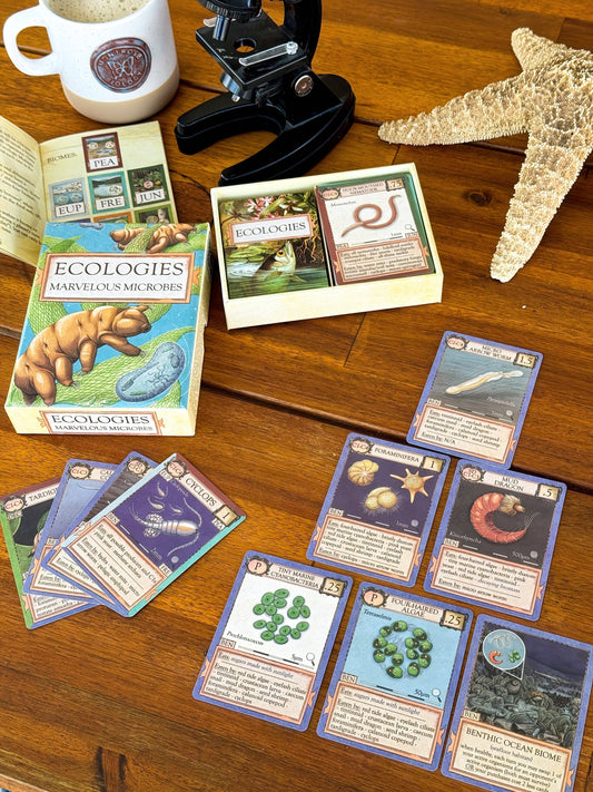 Ecologies: Marvelous Microbes card game with cards and rulebook arranged on a wooden table.