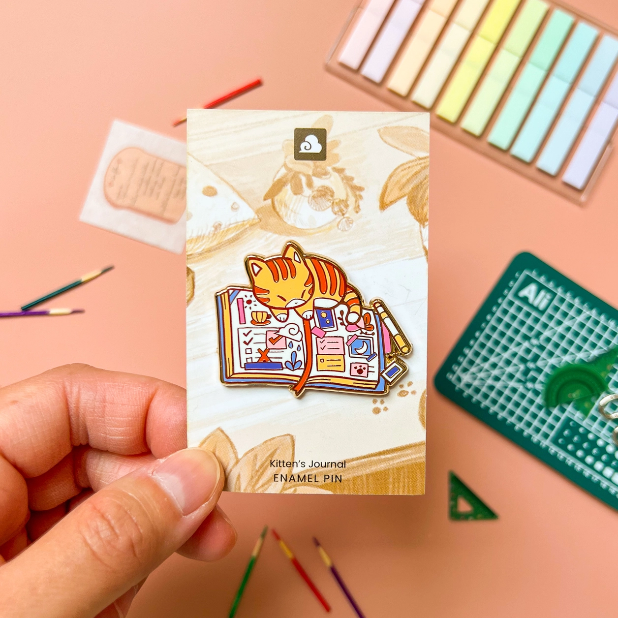 Kittens Journal Enamel Pin featuring an orange cat on an open journal with art supplies, held in a hand.