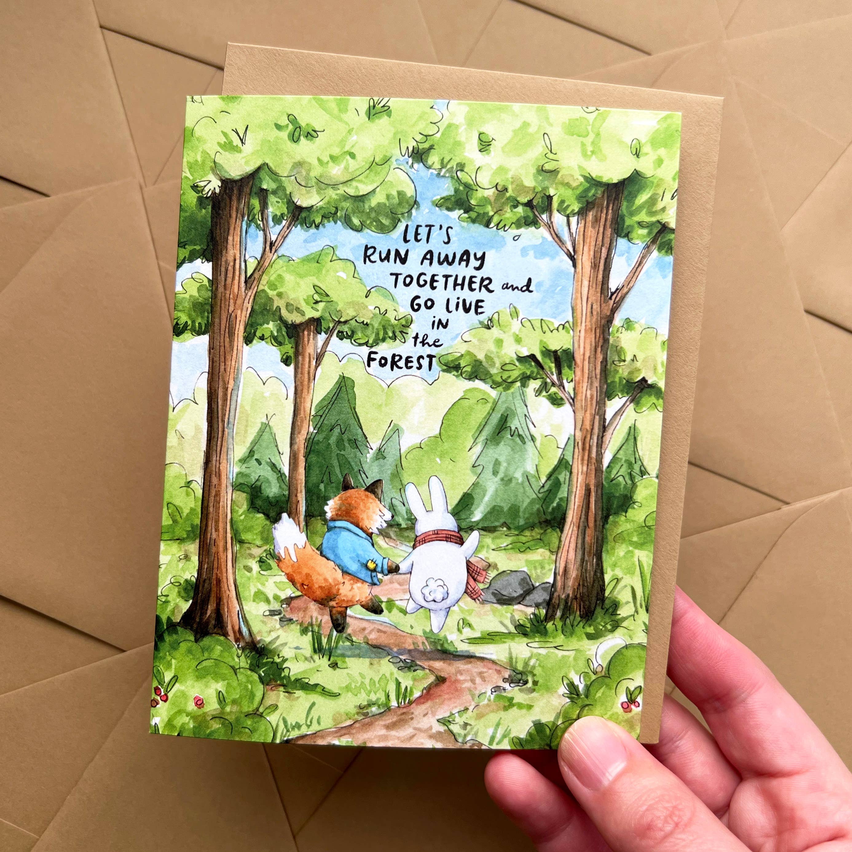 Watercolor greeting card featuring a fox and rabbit holding hands with the text Lets Run Away Together And Go Live In The Forest.