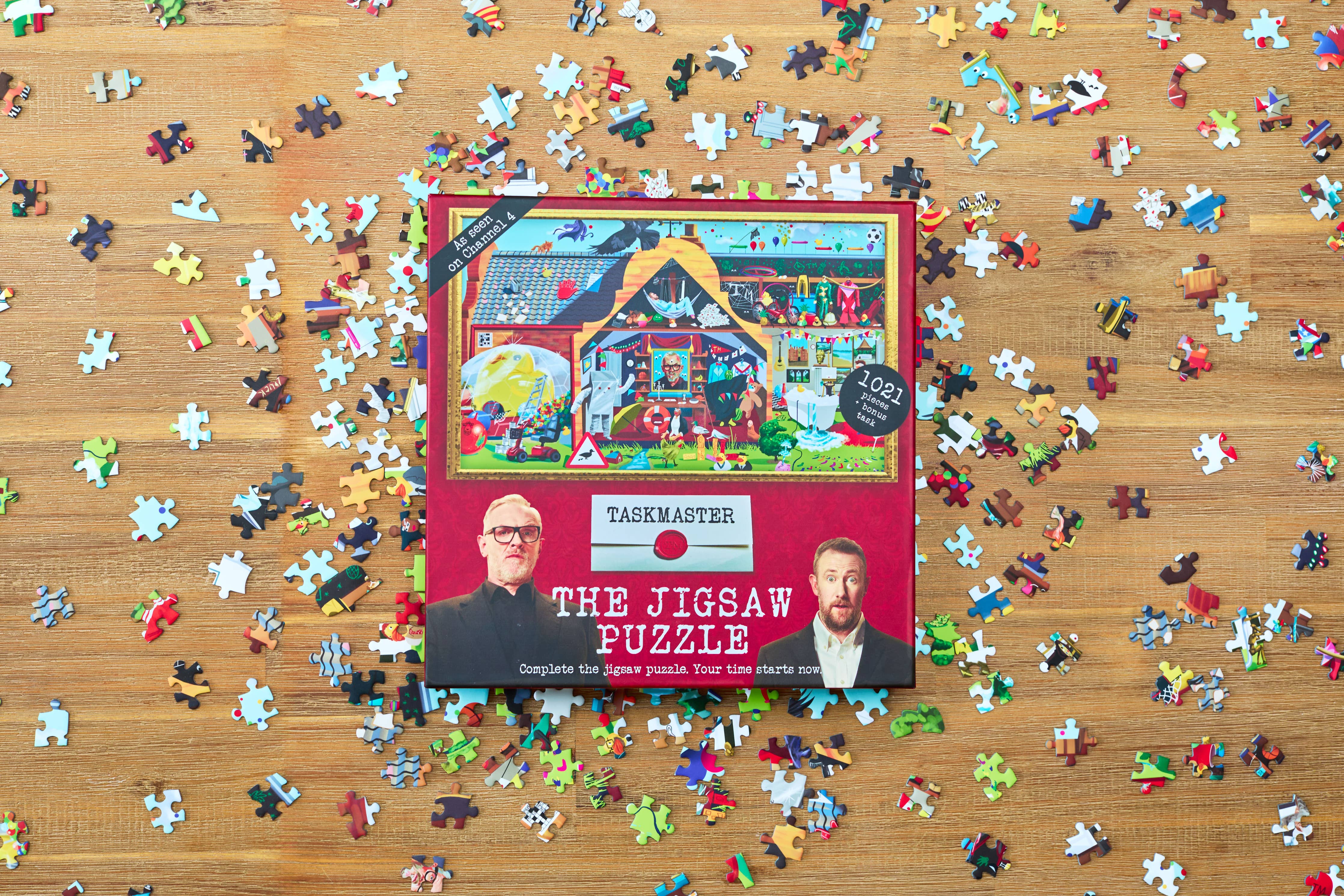 Taskmaster Jigsaw Puzzle (1021 pieces, Secret Task) box with loose puzzle pieces on a wooden table.