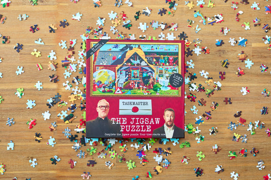 Taskmaster Jigsaw Puzzle (1021 pieces, Secret Task) box with loose puzzle pieces on a wooden table.