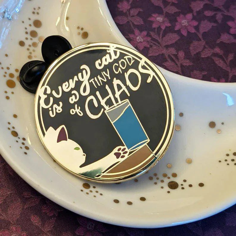 Chaos Cat hard enamel pin, black and gold: cat toppling glass with Every cat is a tiny god of chaos text.
