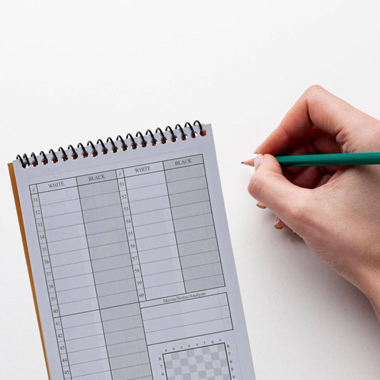 WE Games Ultimate Chess Players Scorebook with a hand holding a pencil above it on a white surface.