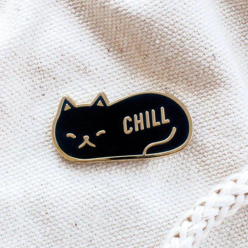 Gold plated enamel pin shaped like a relaxed cat with CHILL on its body, shown on cream fabric.
