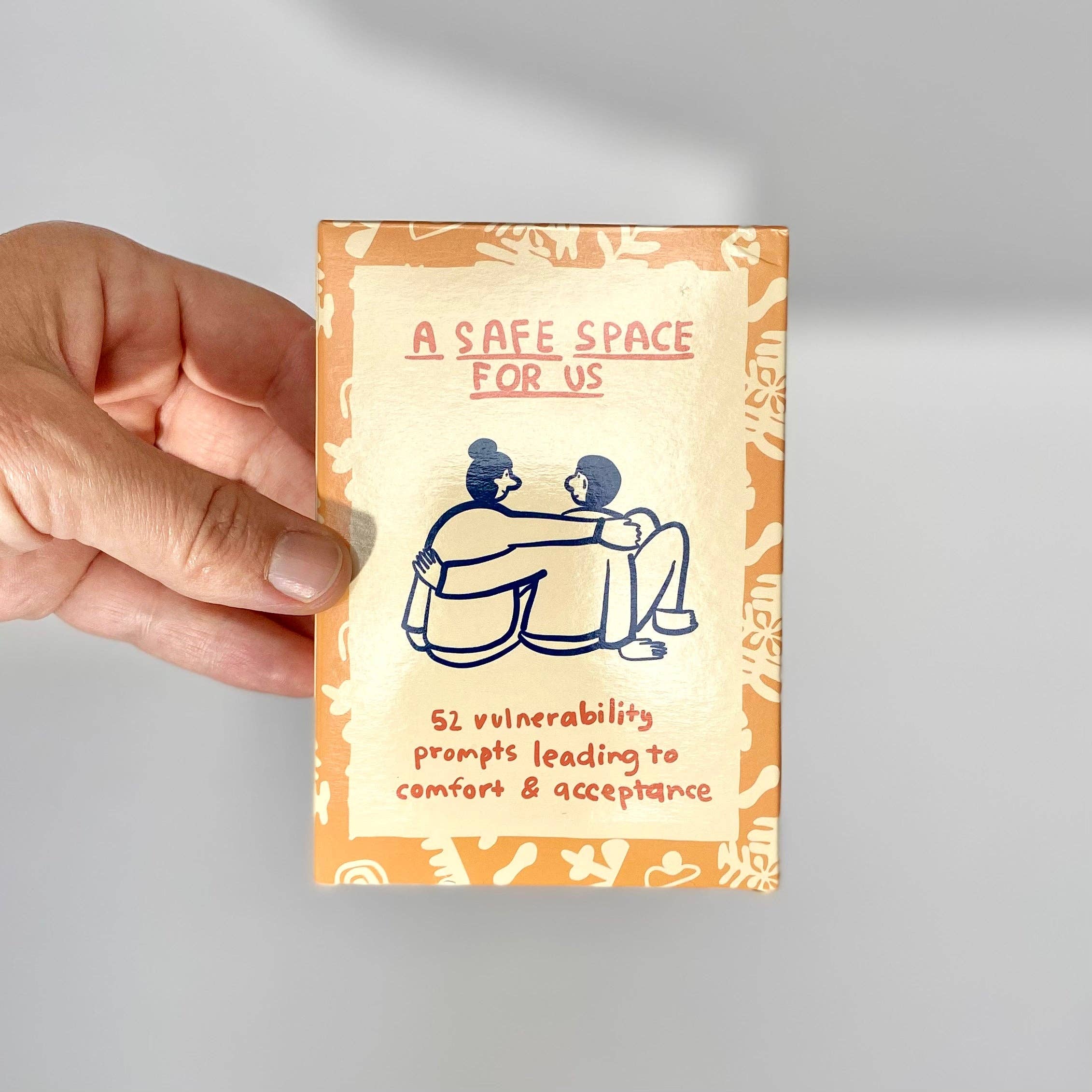 A hand holds the A Safe Space For Us Deck box for couples, featuring an illustration of two people embracing.