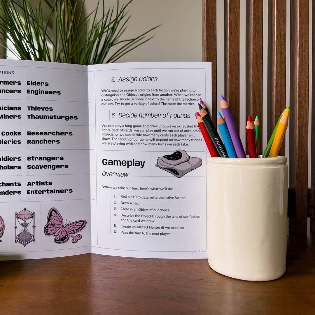 Open rulebook for The Peddler’s Prize inventory building game with a cup of colored pencils.