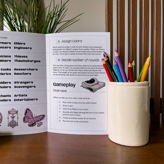 Open rulebook for The Peddler’s Prize inventory building game with a cup of colored pencils.