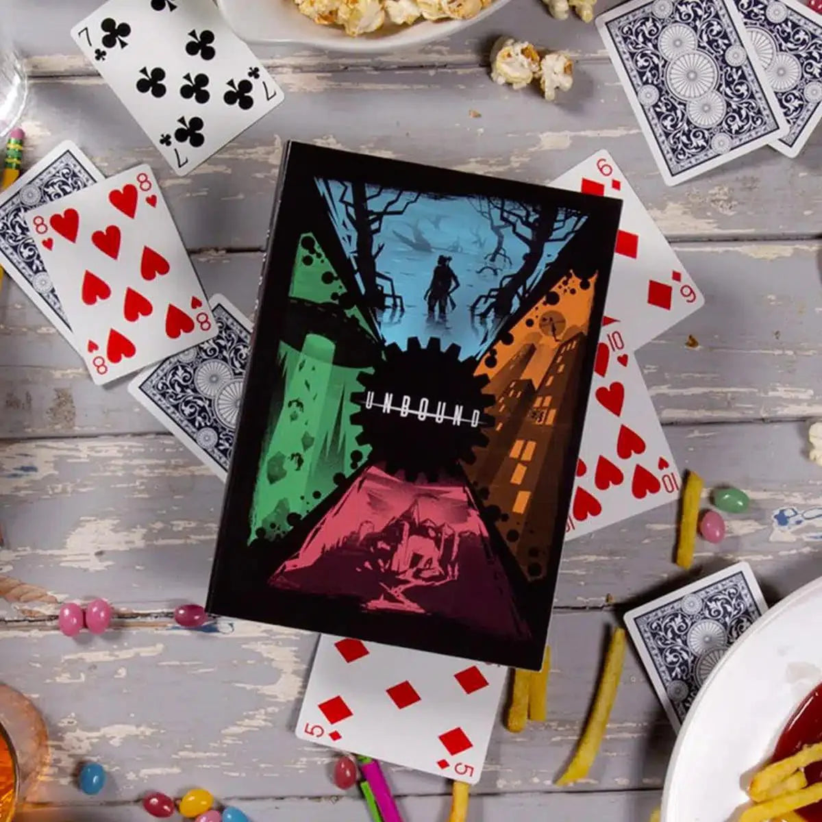 The Unbound RPG game with playing cards, popcorn, fries, and candy on a wooden table.