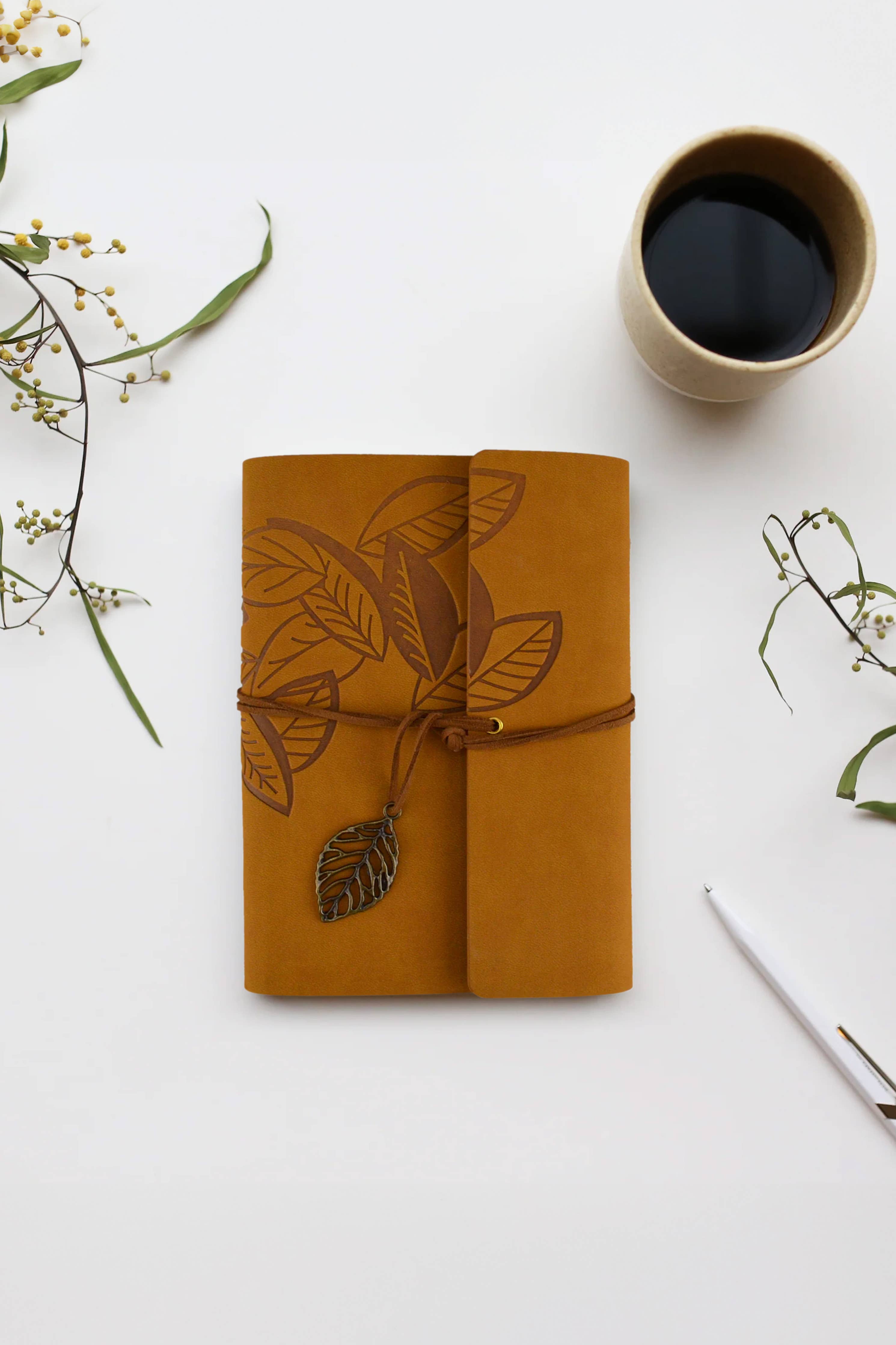 Journal Leaf - Brown: bound notebook with embossed leaves, twine tie, and leaf charm next to coffee cup and pen on white surface.