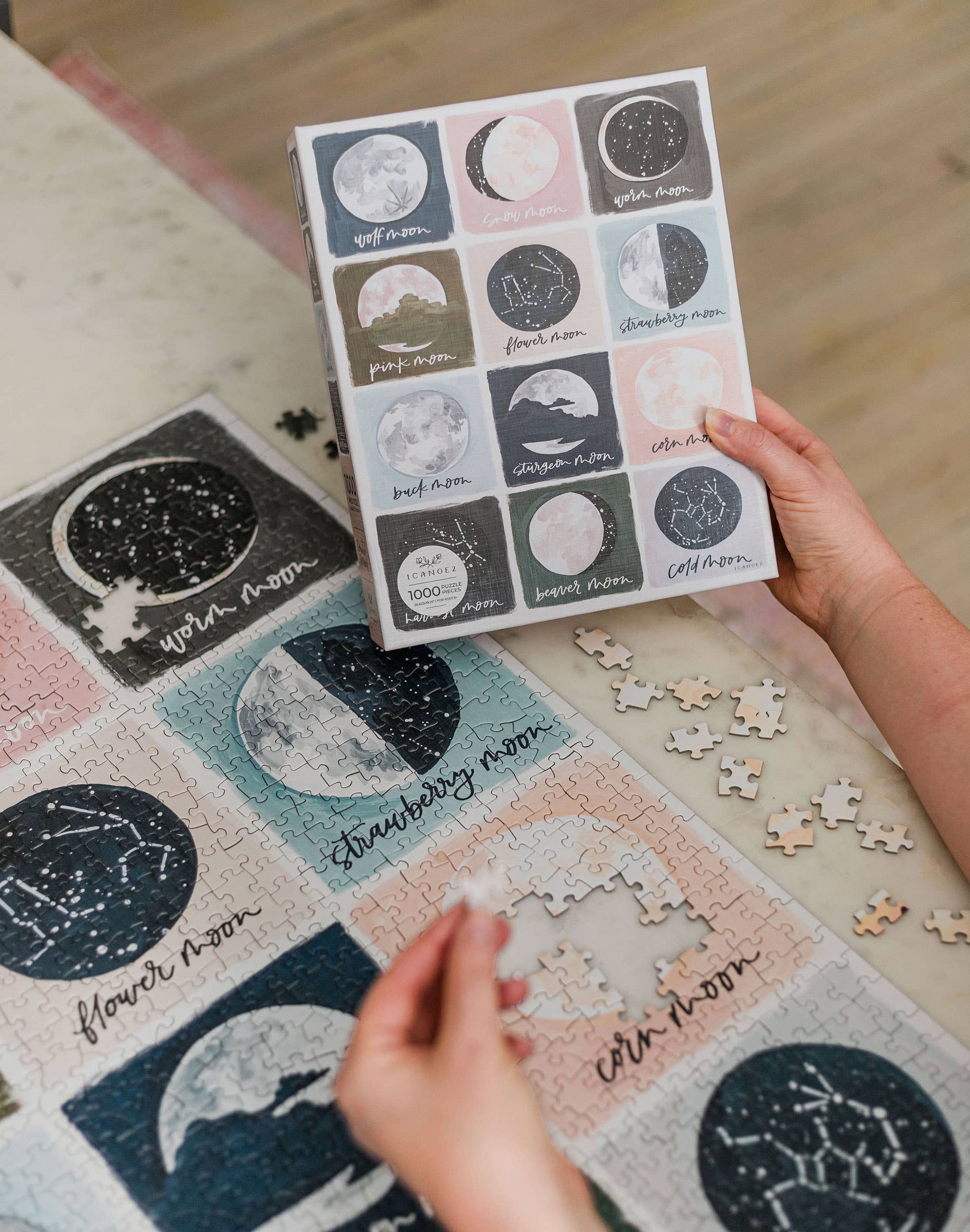 Painted Moons 1,000 piece jigsaw puzzle being assembled; person holds box showing moon phases.