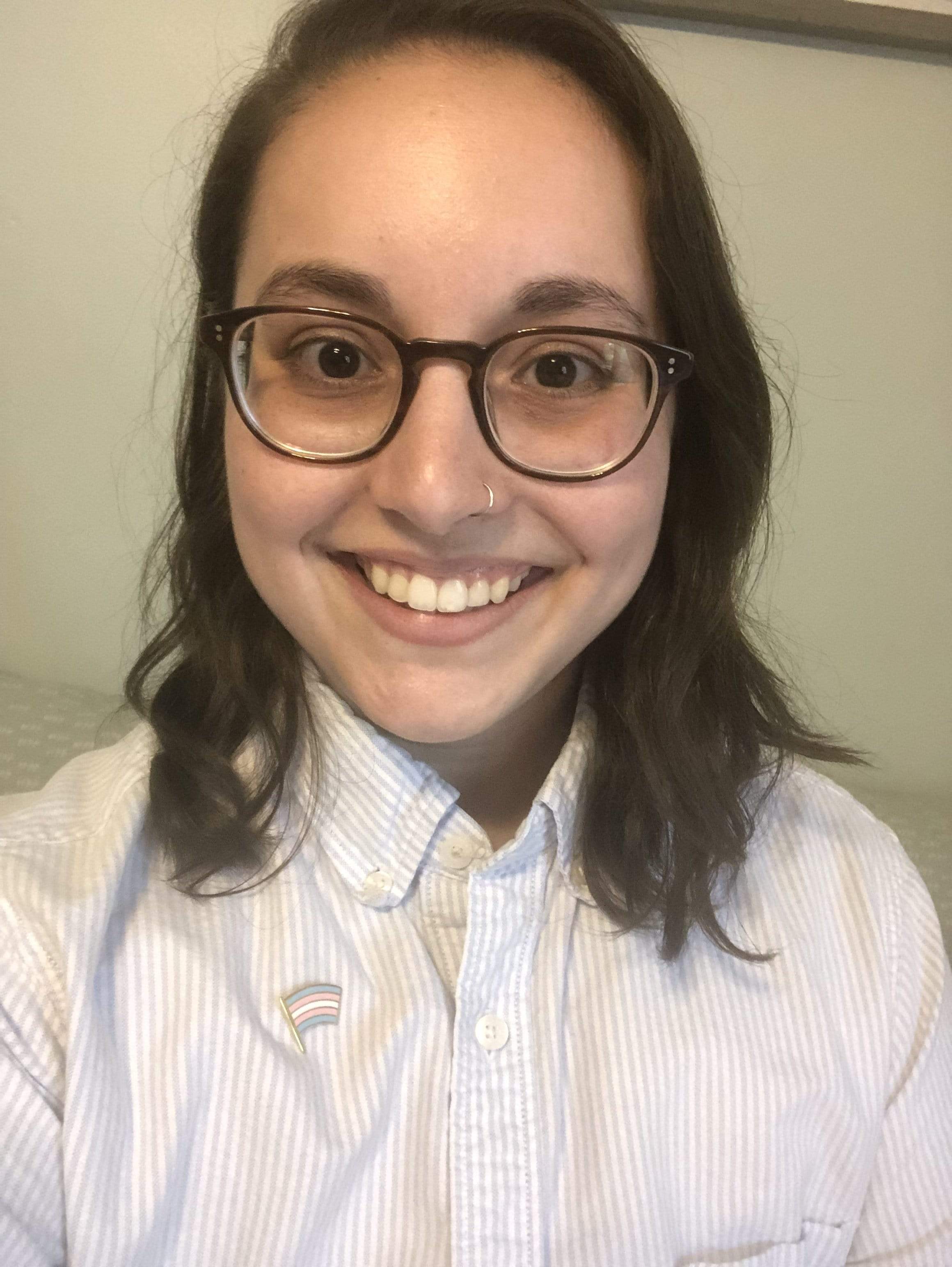 Person wearing glasses, nose ring, striped shirt, and a Trans Pride Flag pin.