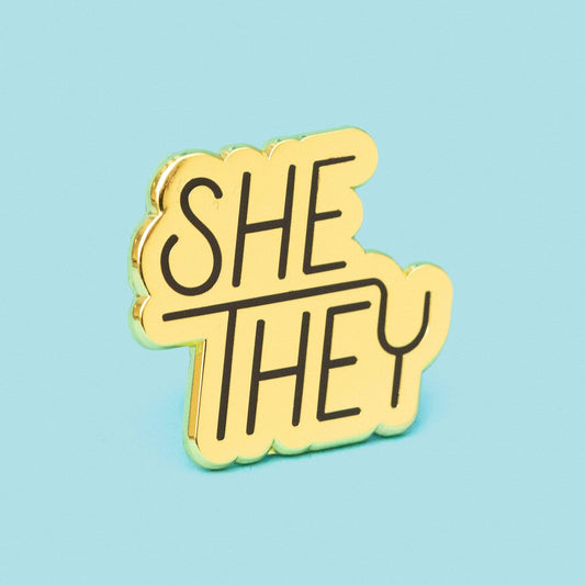 Gold enamel pronoun pin with SHE THEY in black text on a light blue background.