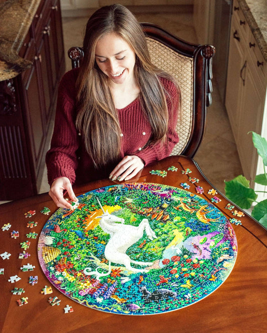 Unicorn Garden 500 Piece Round Puzzle being assembled on a wooden table by a woman in a red sweater.