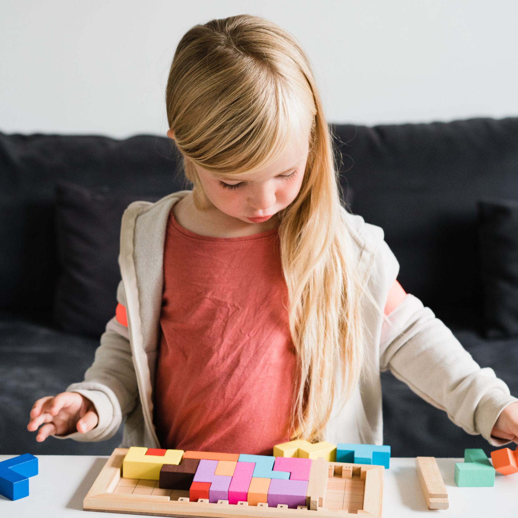Katamino Family puzzle game being played at a white table. Young girl with long blonde hair interacts with the game pieces.