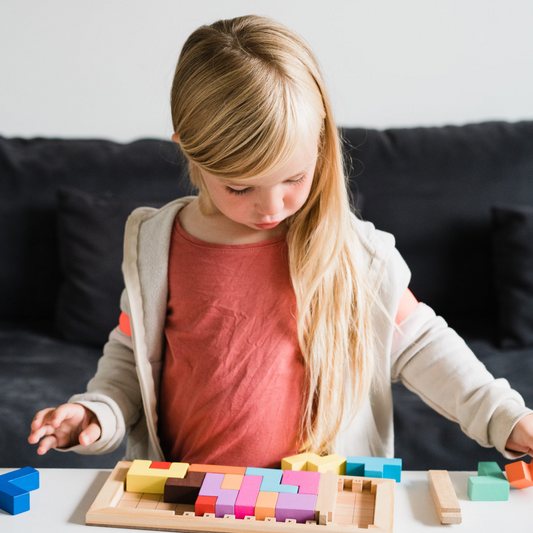 Katamino Family puzzle game being played at a white table. Young girl with long blonde hair interacts with the game pieces.