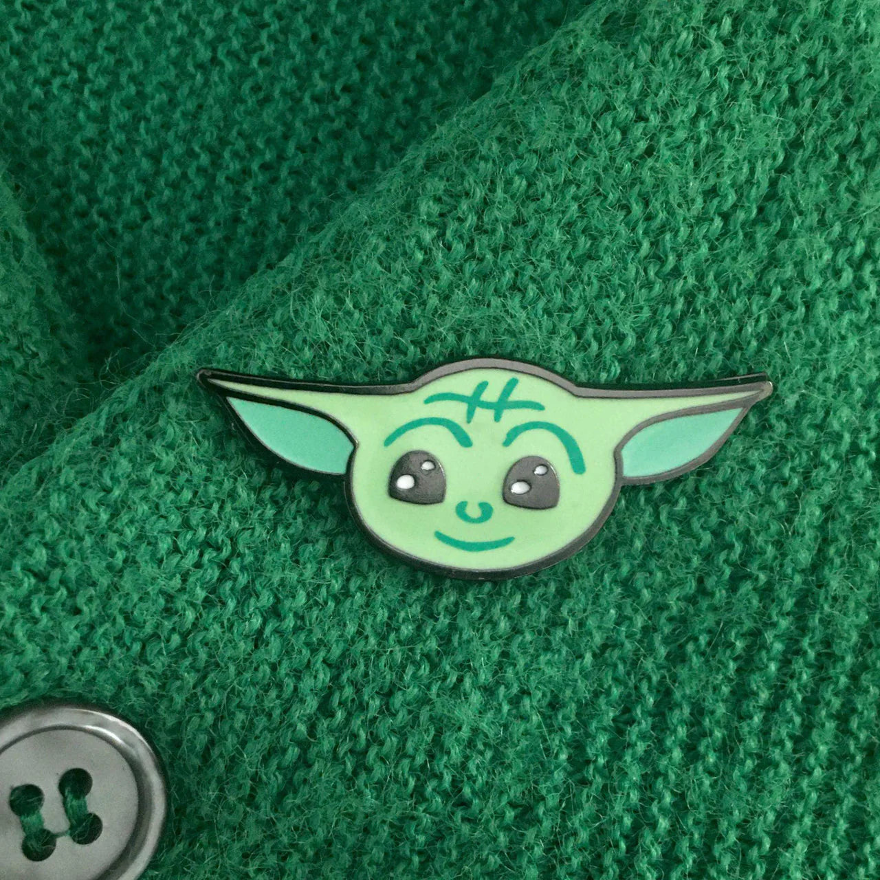 Baby Yoda enamel pin inspired by Star Wars shown on green fabric next to a silver button.