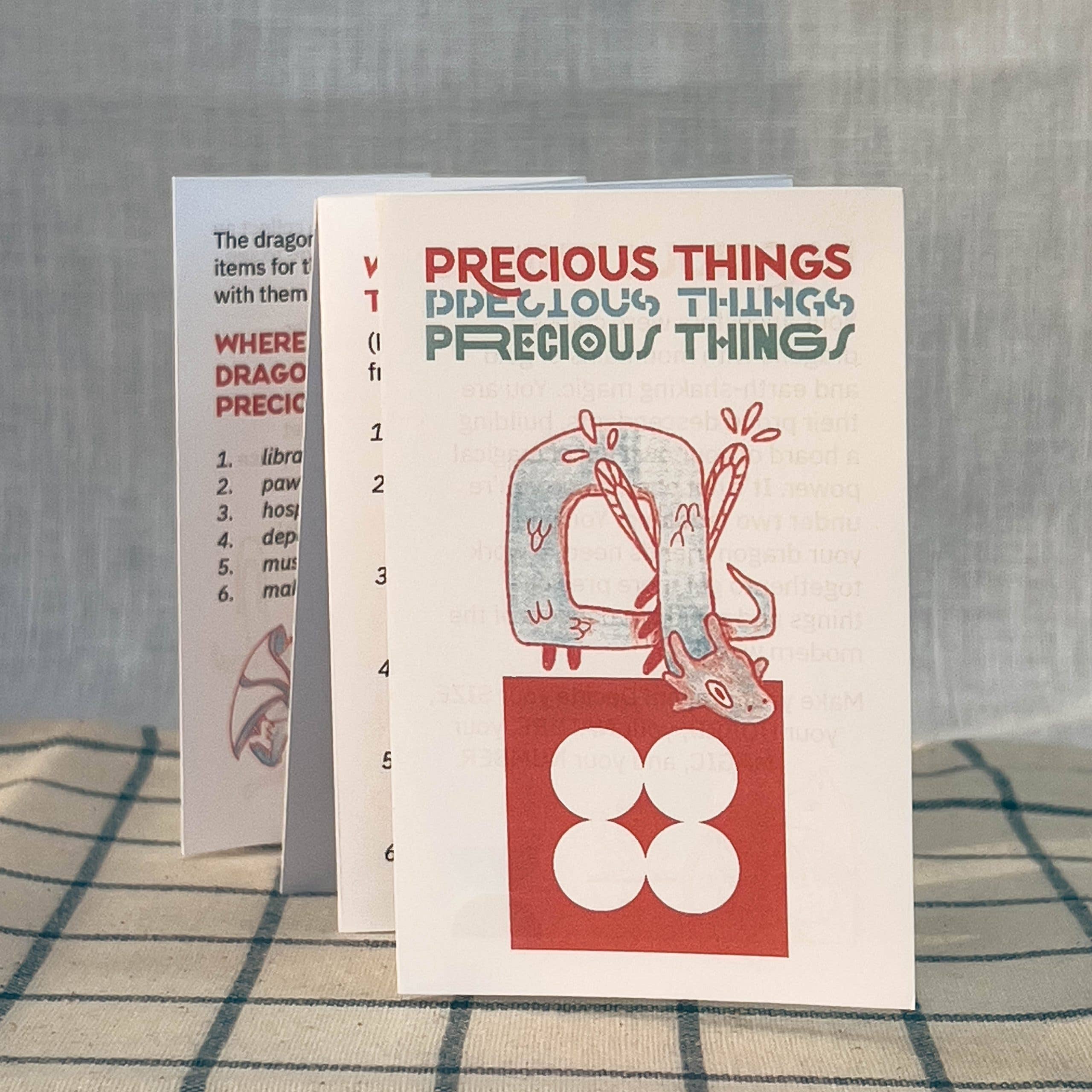 Precious Things, a folded one-page TTRPG zine featuring a tiny dragon, a red square with white circles, and locations.