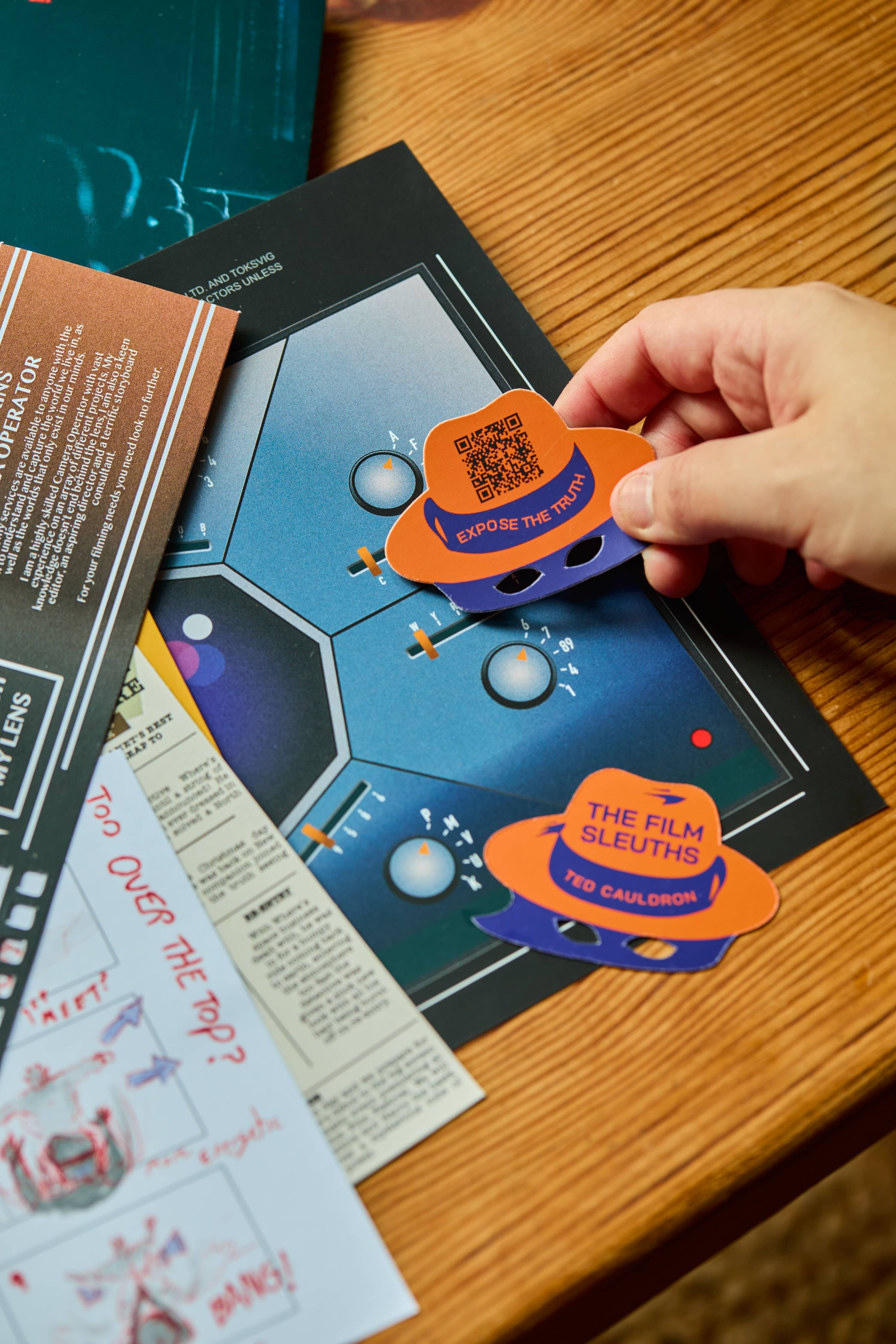 Hand holds orange token labeled THE FILM SLEUTHS over “The Betrayal” Escape Room Puzzle Board Game on table.