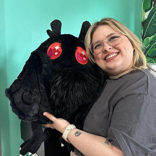 Person with glasses holds a large Squishable Baby Mothman plush with big red eyes in a brightly lit room.