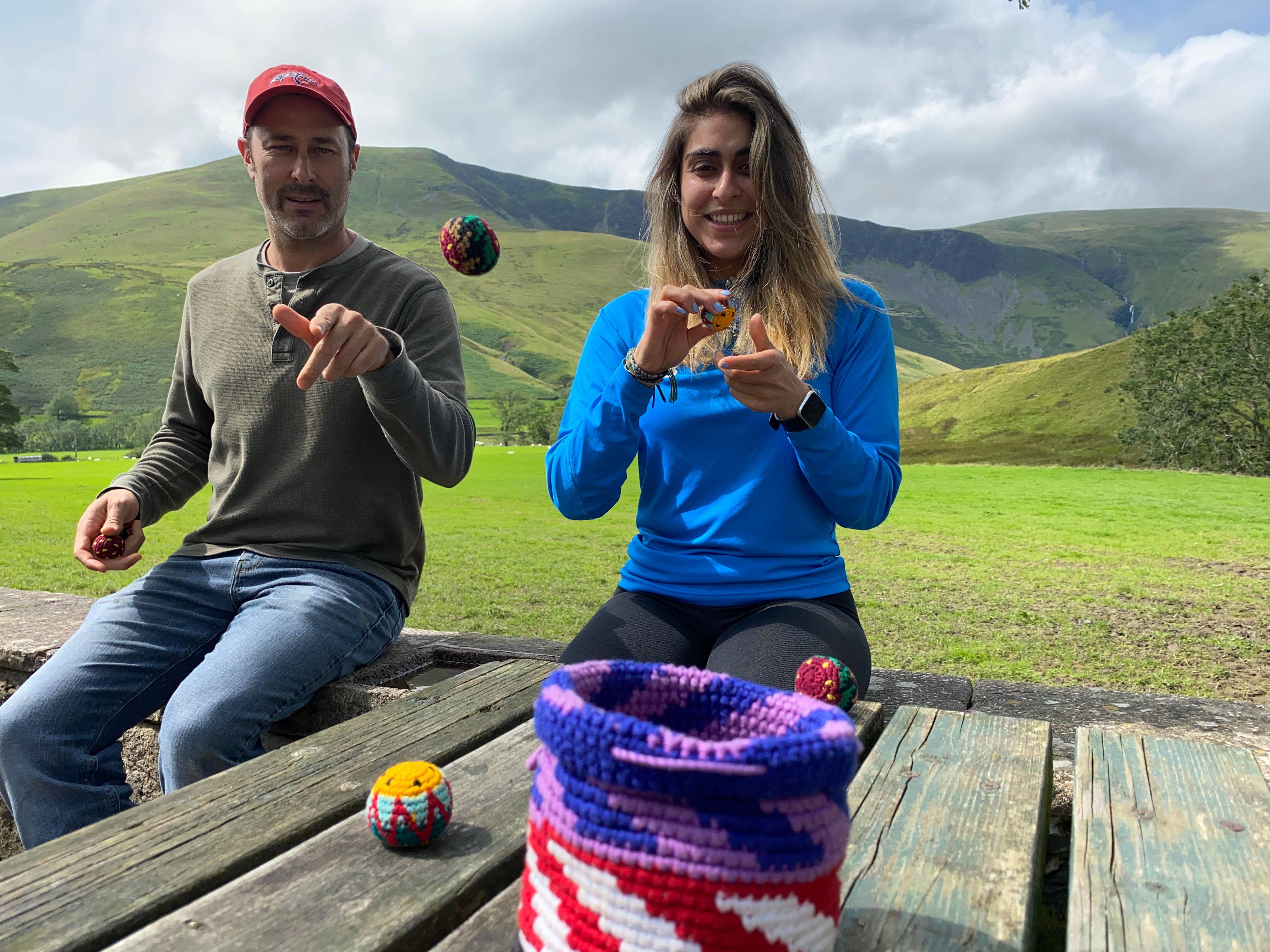 Two people at a picnic table outdoors toss Canastaball Hacky Sacks. Green hills are visible in the background.