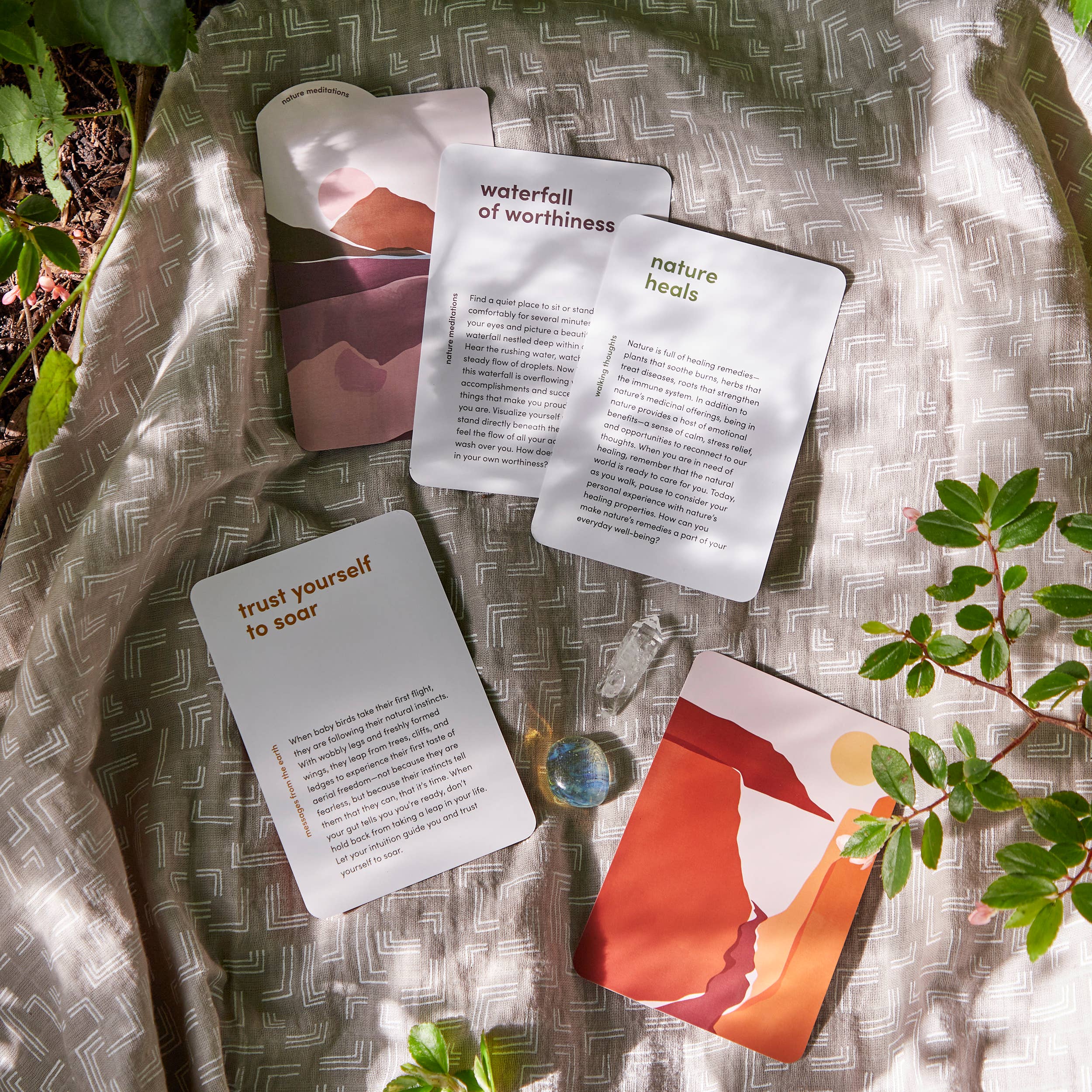 The Nature Meditations Deck and a glass sphere on patterned cloth with green leaves in sunlight.