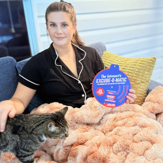 Woman in pajamas holding The Introverts Excuse-o-Matic, with a cat nearby.