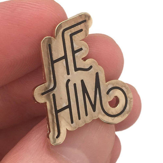 Gold Pronoun Pin with He/Him in black lettering held by a hand.