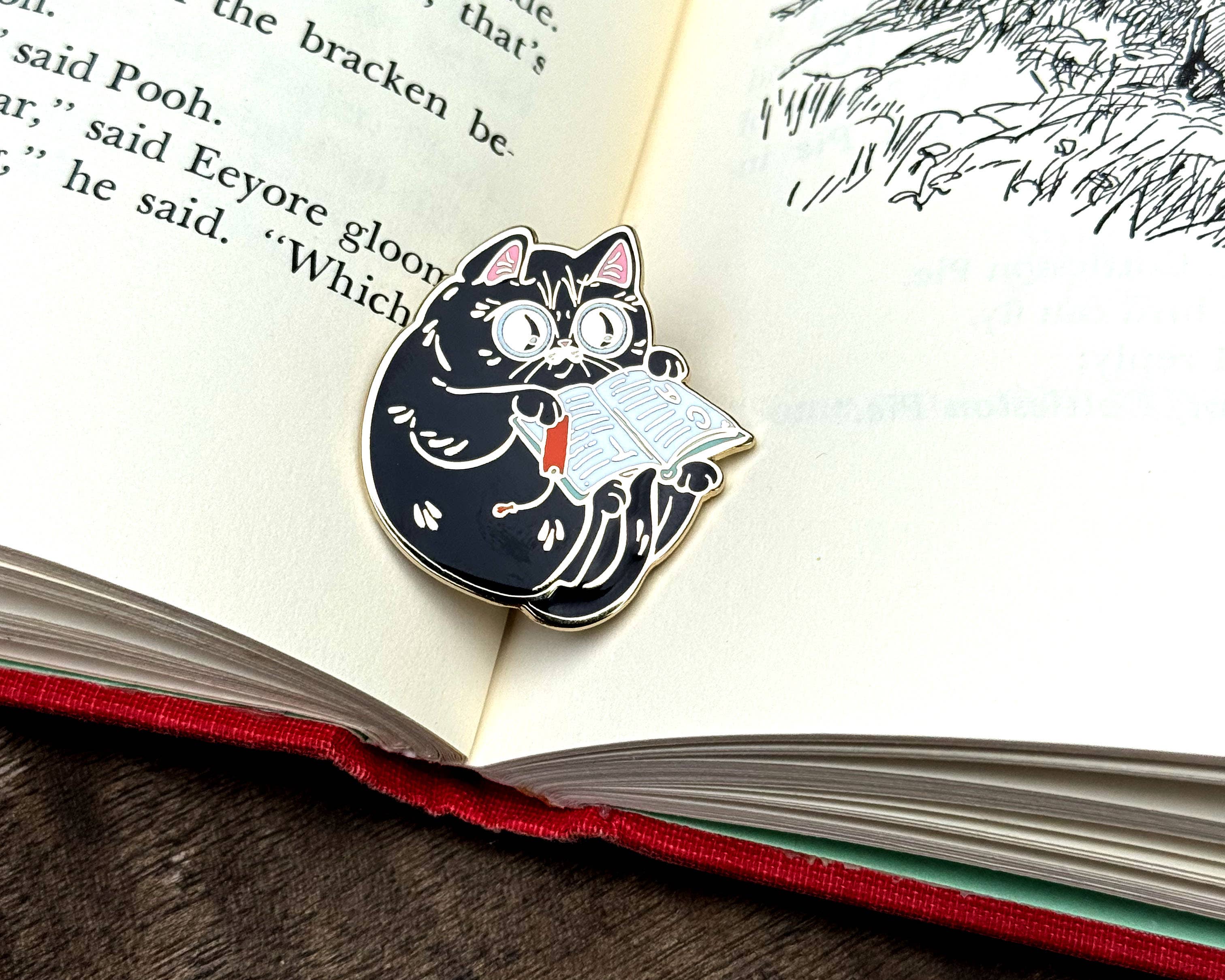 Enamel pin of a wide-eyed black cat reading an open book with a red bookmark.