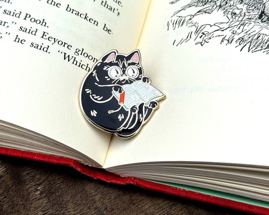 Enamel pin of a wide-eyed black cat reading an open book with a red bookmark.