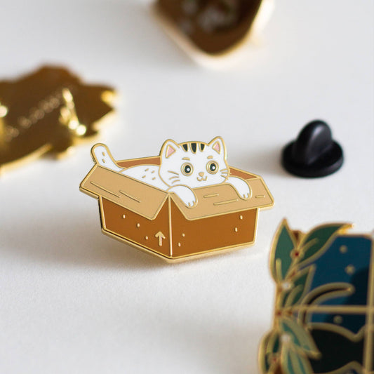 Cute cat enamel pin peeking from a box, perfect for any cat lover gift, displayed among other pins on a white surface.