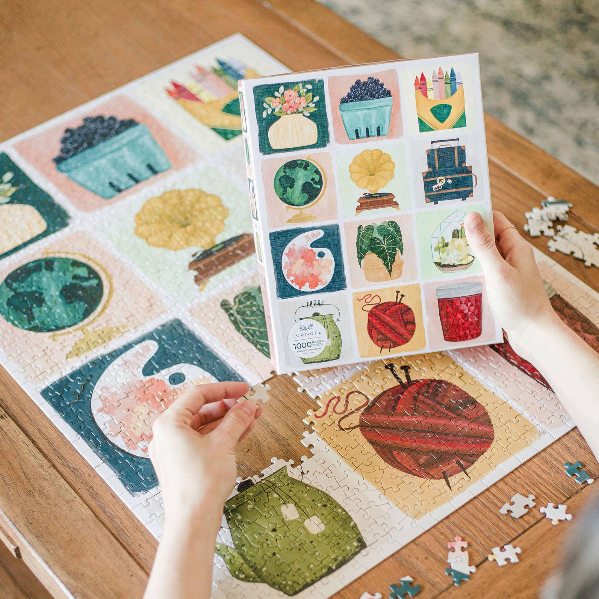 Simple Joys Puzzle, 1,000 pieces, with hand-painted plants, books, yarn, and globes being assembled by a person.