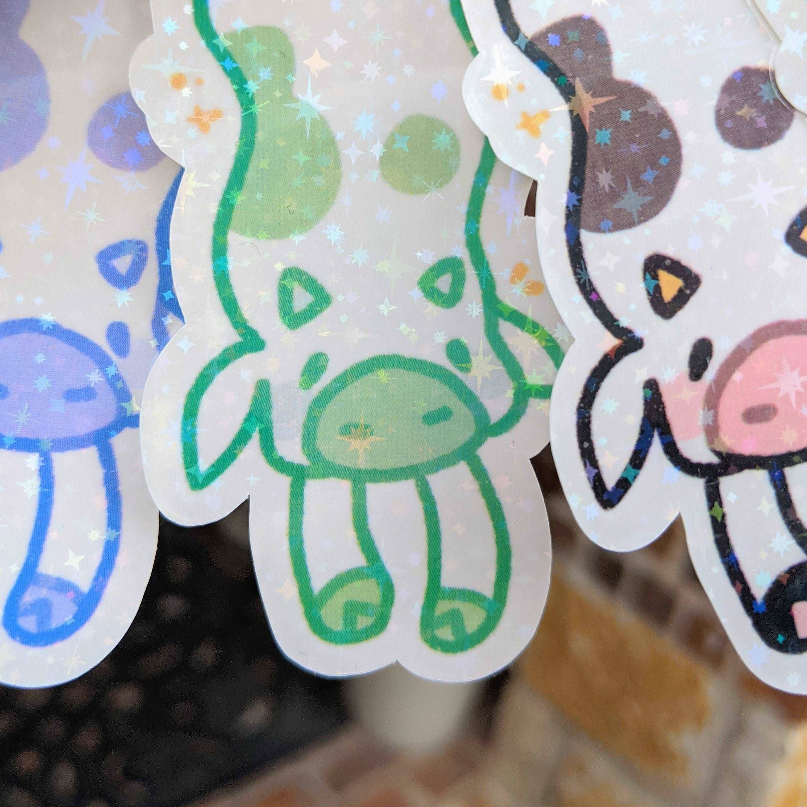 Set of three sparkly long cow stickers, each outlined in blue, green, or black with holographic stars on translucent backing.