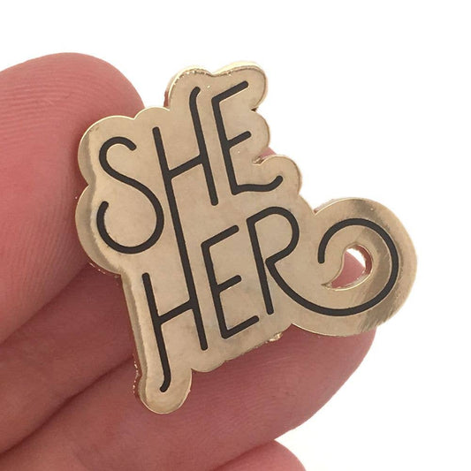 She/Her Pronoun Pin, gold with bold decorative letters, held in a hand.