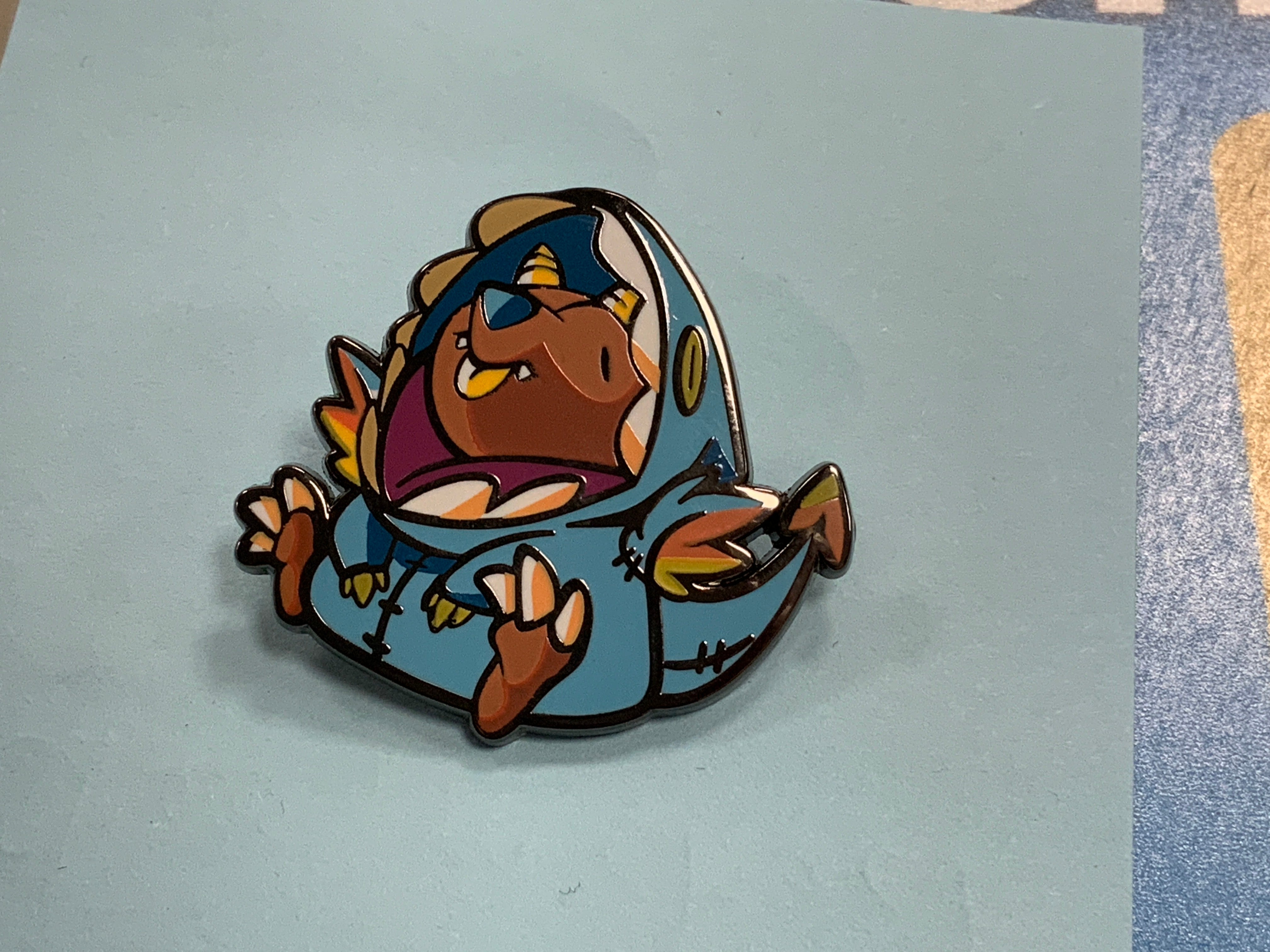 Heliana Monster Hunting Rakitten enamel pin in a blue dinosaur costume.