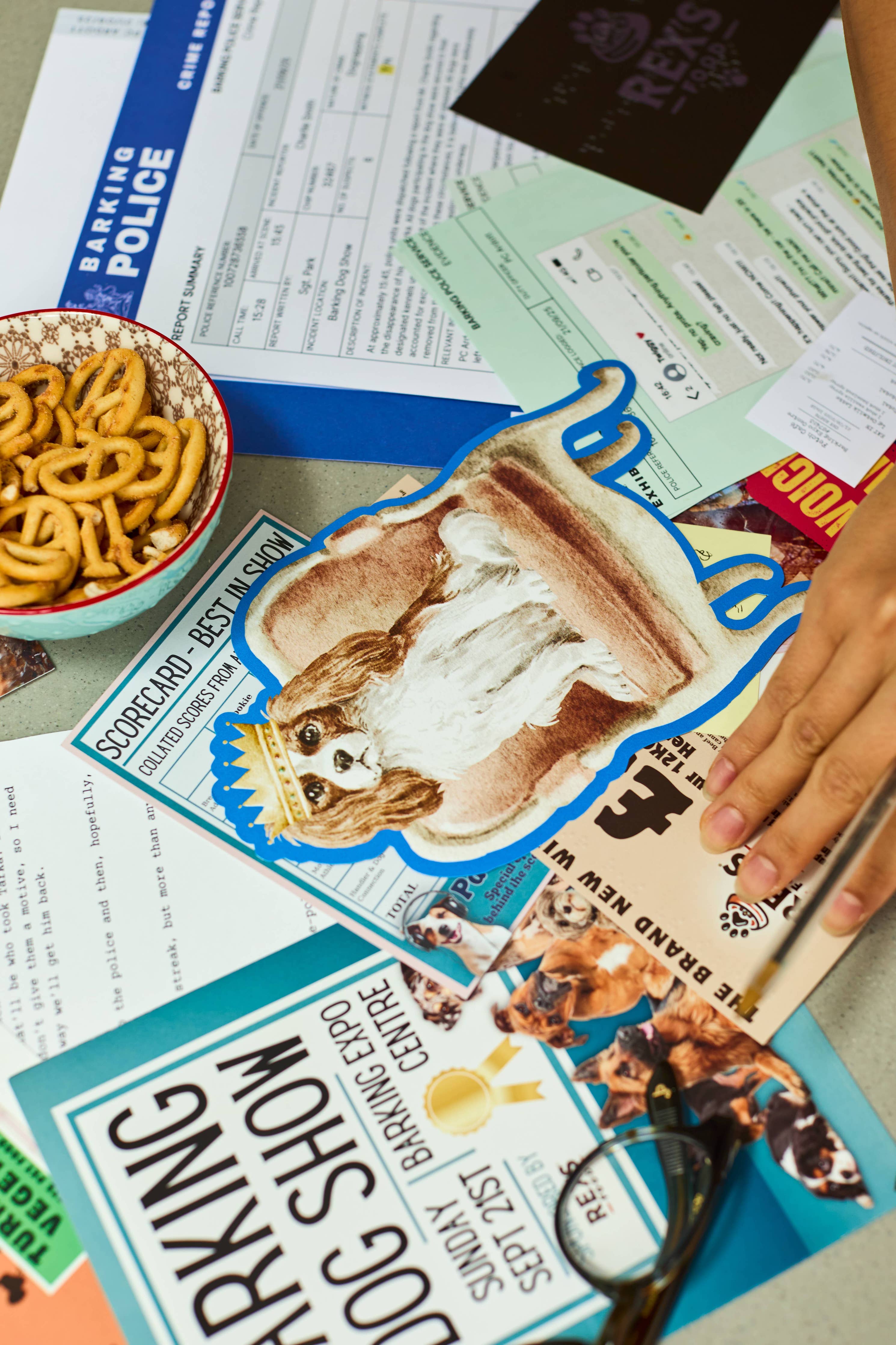 Hand touching dog show papers on table with snacks, tickets, and Cold Case: Who Let The Dogs Out? magazine.