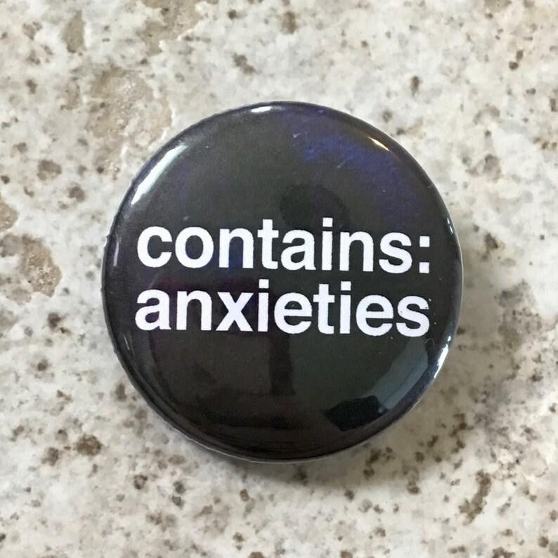 Round black 1.25 pin with bold white text reading Contains Anxieties on a speckled background.