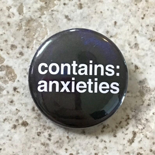 Round black 1.25 pin with bold white text reading Contains Anxieties on a speckled background.