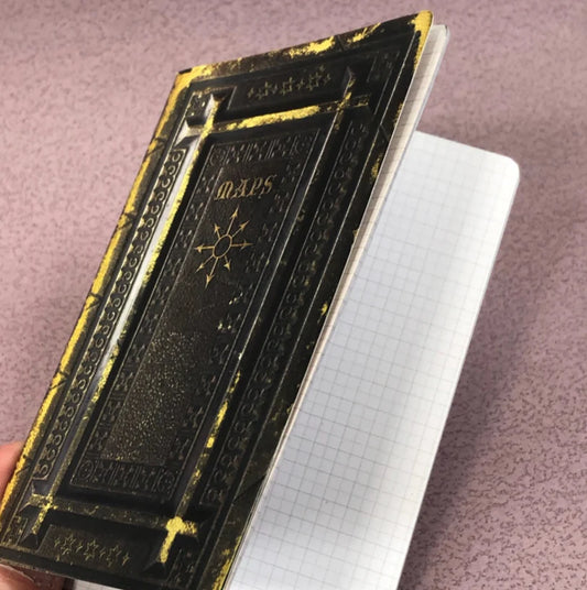 Ancient Tome Cartography Mapping RPG Notebook with black and gold Maps cover and graph paper pages held in a hand.